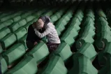 Two women hold one another as they stand amid many coffins arranged in rows.