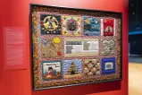 A colourful quilt, framed and hung on a red wall. The quilt includes several images including one of a girl washing a floor, a heart and Indigenous symbols.