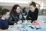 A smiling parent and two children sit at a table, with crafting supplies including markers, scissors, glue and construction paper laid out before them.