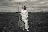 A woman in a long white dress stands in a field holding a bandura, a Ukrainian plucked string folk instrument.