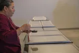 Framed documents sit on a table as Elder Hazel Copenace smudges them, holding a smudge bowl in her left hand and an eagle feather in her right.