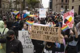 A group of people crowd into a city street holding signs and rainbow flags. The sign in the foreground reads, “To protect all kids we must protect trans and queer kids.”