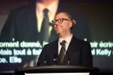 A white man with blond hair standing before a podium and in front of a screen is smiling off to the corner of the camera. He is wearing a black suit with a dress shirt and dark gray necktie.
