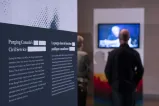 An exhibit panel with the text “Purging Canada’s Civil Service” is on the left. Two people in the background on the right watch a video. 