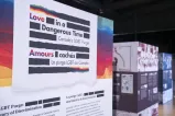 A large sign is made up of thick black horizontal lines and the English and French words “Love in a Dangerous Time: Canada’s LGBT Purge” and “Amours cachés : La purge LGBT au Canada” are distributed within the lines. The top and bottom lines are a mix of black and rainbow colours.
