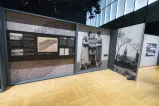 A large exhibit panel with text and historical images is on the left. On the right is a large black-and-white photograph from the 1910s of a man in a military uniform flanked by two women in dresses. A photograph of a large building can be seen further to the right.