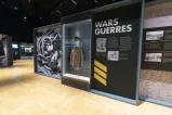 A series of exhibition panels feature historical images of military personnel. The word “Wars” is on the top of one panel. A display case located between two panels contains a First World War military uniform.