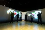 A group of people wave their arms in front of a darkened wall. Colours, shapes, and words appear on the wall where their hands are.