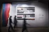 A small group of people walk by a large sign on a wall, made up of thick black horizontal lines with the English and French words “Love in a Dangerous Time: Canada’s LGBT Purge” and “Amours cachés : La purge LGBT au Canada” distributed within the lines. The top and bottom lines are a mix of black and rainbow colours. Rainbow colours cover the left edge of the wall.