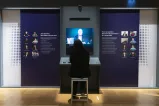 A person with their back to us watches a video. Small portraits of people flank the video monitor on the left and right.