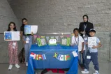Four students and an adult stand next to a table with flags from countries around the world. Three of them are holding up an open book.