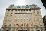 A large historic hotel building with a beige stone façade, green copper roof, and ornate architectural details. A Canadian flag is flying at the top of the building and several flags are displayed at the entrance below.