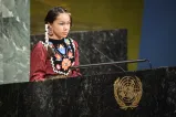 An Indigenous girl is wearing traditional regalia and beaded earrings and her hair is in braids. She is speaking at a podium featuring a logo of the United Nations.