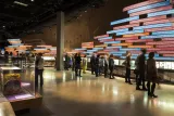 A large crowd in a Museum gallery. Most people are looking at a display on the wall of the gallery. The display consists of a series of pictures and of long rectangular text panels placed horizontally.
