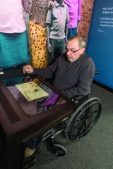 An older man sits in a wheelchair in front of a counter with a large touchscreen.