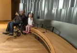 An older man sits in a wheelchair beside a young girl sitting on a bench. The man is pointing up, and both he and the young girl are looking up and to the viewer’s left.