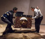Luke Marston (left) and another person lift the Bentwood Box from a buffalo hide, which lies on the floor underneath.