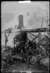 A black and white image of nine men in front of a logging camp. Three are standing while the rest are sitting.
