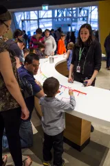 Museum visitors of all ages are writing on cards and talking to one another around a curved table. 