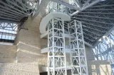 The interior of a large open spaced part of the Museum with two elevator shafts in the centre of the image, and a limestone wall.
