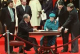 Queen Elizabeth II sitting at a table and signing a document. A man is also sitting to her right, and others are standing behind them around the table.