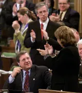 In the House of Commons, a man is sitting while his fellow politicians are standing and applauding.