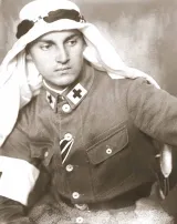 White man in an army uniform with a black cross at the edge of his collar wears a keffiyeh, white headdress worn in the Middle East.