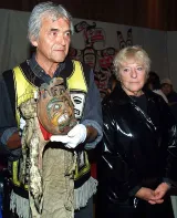 Bill Cranmer, an older man with grey hair, holds a cloth and fur headdress. It features a painted round face carved from wood. To the right of Cranmer is Aube Elléouët, an older woman with short hair. She is wearing a dark jacket.