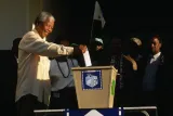 An elderly Nelson Mandela smiles as he reaches out with his right arm to put a ballot in a large metal ballot box.