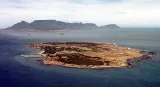 An aerial view of Robben Island. The island is very flat and roughly oval in shape, with a little under half its surface covered with trees. An airfield is visible on the right side of the island. In the distant background behind the island, the mainland can be seen, including the buildings of a city. Rising above the city are large mountains. 
