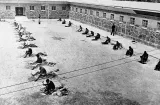 A black-and-white image of two rows of men in a courtyard. The men are sitting and breaking stones with small hammers.