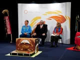 Two men and a woman sit in front of a white background decorated with stylized flames. To their right is a vertical banner reading "Truth & Reconciliation Commission of Canada", and in front of them is a rectangular carved wooden box. On the side facing the camera is a round, weeping face.