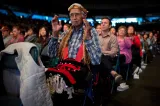 An elderly man wearing a checkered shirt and traditional Indigenous accessories is sitting in a crown. His arms are bent at the elbows and his hands are up facing each other.