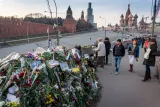 A huge pile of flowers, photographs and messages sits on a bridge beside a road. People are walking over the bridge. Some are looking at the pile of mementos. One woman is taking a photograph with her tablet. In the background is a red concrete wall (Red Square in Moscow) and a church with curvy, domed spires (St. Basil’s Cathedral).