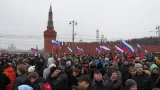 A crowd of people marches holding the red, white and blue striped flags of Russia. Behind is a red concrete wall that ends in a spire.