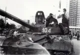 Two men stand on a parked military tank, talking. There are bouquets of flowers strewn across the tank. Behind are people milling in front of a tall building. The photo is black and white.