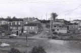 A black and white image of about a dozen houses on a hill. In the middle of the image, a dirt road travels up the hill between the houses.