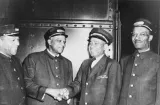 A black and white photo of four men in train porter uniforms. All of the men are smiling, and the two men in the middle appear to be shaking hands.