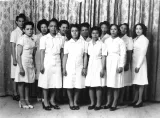 A black and white image of Viola Desmond with 11 other women. All of the women are wearing plain white dresses and are standing in two rows, posing for the camera.
