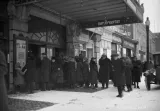 A black and white image of a crowd in front of a theatre entrance. The theatre is named “Metropolitan”. Everyone is wearing long old-fashioned coats, and some are wearing hats. An antique car is parked on the street.