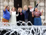 Six women are standing in front of what appears to be the entrance of a large stone building. They are all smiling and holding up one or two hands making the peace sign.