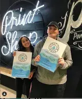 A young woman and man hold up posters that promote a fashion show. In the background are bright neon lights.