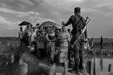 A soldier with a rifle on his back blocks a group of Rohingya before they cross a makeshift bridge. His left hand is raised, gesturing for them to stop.