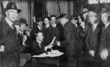A group of men crowd around a man at a desk.