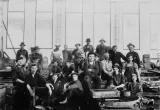 A group of men in working clothes sitting and standing in a factory.
