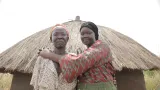 Two women are standing in front of a hut with a straw roof, looking at the camera. The younger woman on the right is smiling and embracing the one on the left.