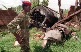 A soldier carrying a rifle and wearing a red beret and camouflage uniform stands in front of the wreckage of a plane among trees.