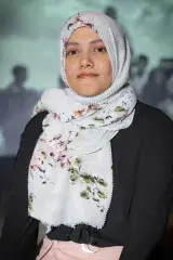 A portrait of a young woman with dark eyes wearing a black long-sleeved top. Her hair is covered with a mostly-white head scarf that has a green and pink floral pattern on it.
