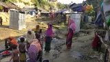 Women and children appear to be washing items in a mostly dried-up stream surrounded by mud. Makeshift camp structures are clustered in the background.