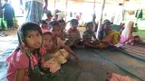 A group of children wearing colourful clothes sit cross-legged on the ground inside a shelter. Adults stand behind them, and there are clothes hanging on walls.